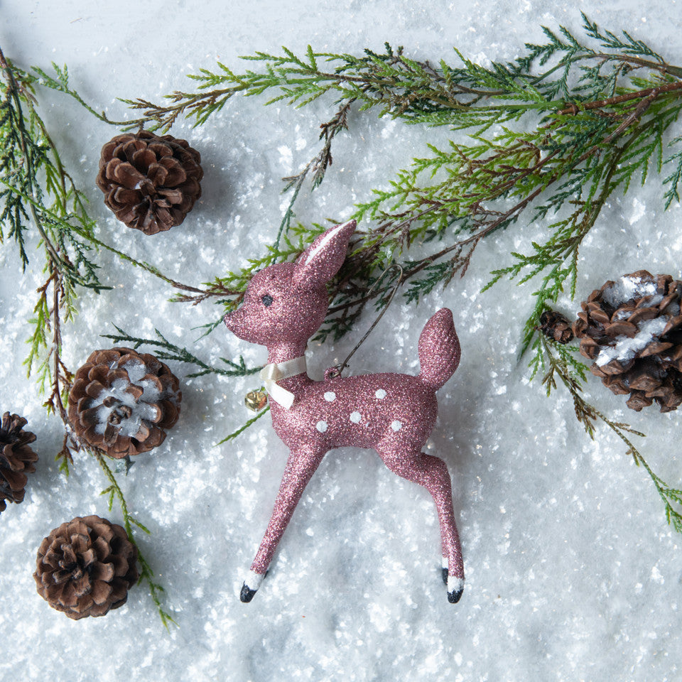 Pink glitter deer ornament with white spots on a snowy background with pine branches and cones.
